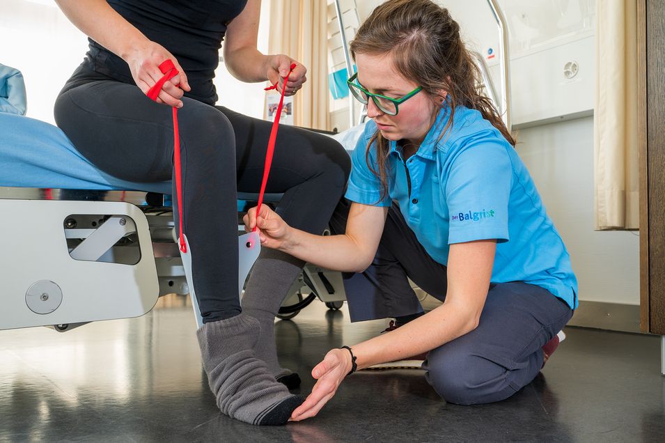 Eine auf einer Liege sitzende Patientin kann dank einer Seil-Vorrichtung ihre Socken selbstständig anziehen.