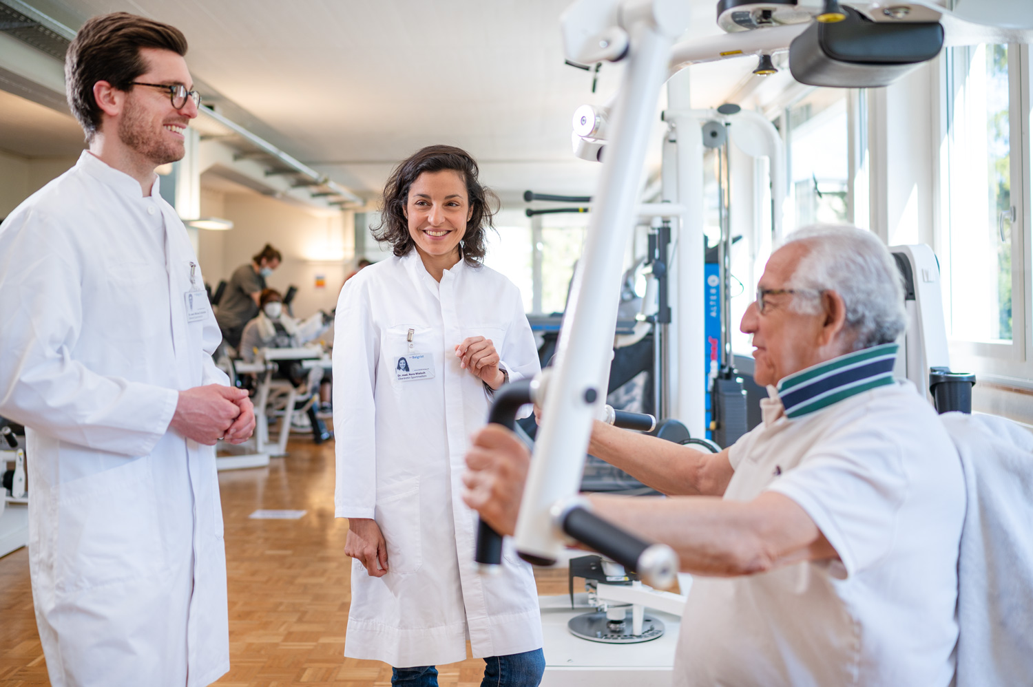 Ein Sportarzt und eine Sportärztin beraten einen Patienten, der an einem Fitnessgerät trainiert.
